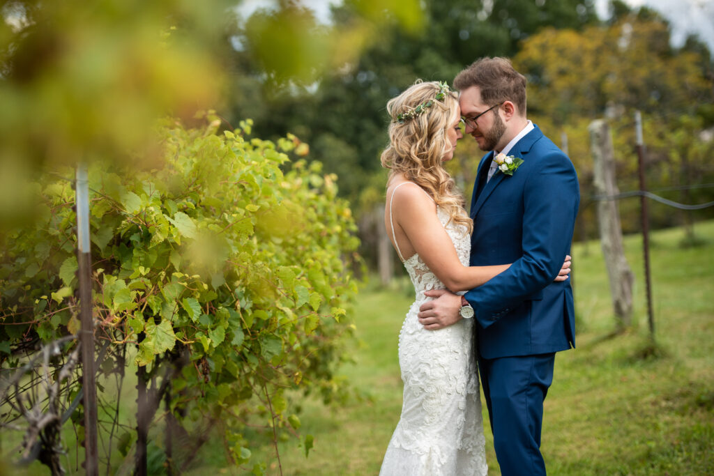 A bride and groom embrace each other amidst grape vines in a vineyard. The couple is holding each other closely, with the groom's hands on the bride's waist.